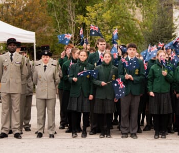A group of school students in green uniforms are posed in a group waving Australian flags. There are a group of adults in French military uniform next to them.