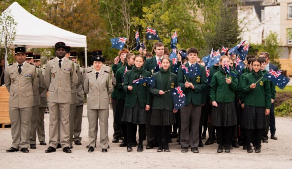 A group of school students in green uniforms are posed in a group waving Australian flags. There are a group of adults in French military uniform next to them.