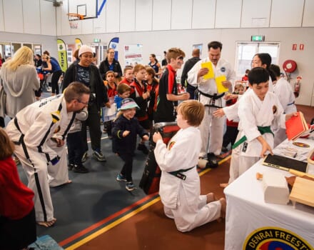 Group of children gathered for a martial arts display, young boy is holding a kick bag for a small child who is about to kick it.