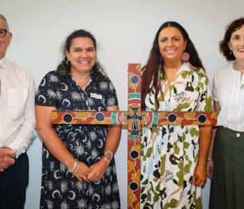 Four people posed smilling with a cross painted in the art style of First Nations People of Australia.