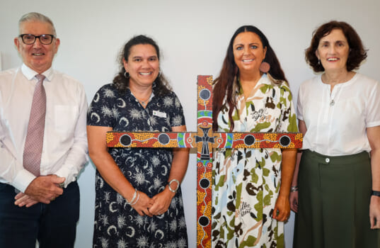 Four people posed smilling with a cross painted in the art style of First Nations People of Australia.