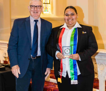 Two people posed together smiling, one is an adult the other is a student in school uniform holding a graduation award