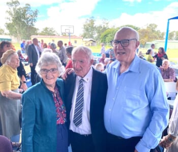 Three elderly people pose together at a large gathering