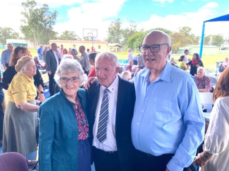 Three elderly people pose together at a large gathering