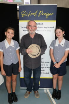 Three people posed in front of a banner. Two are school students in uniform and one is a man holding a hat.