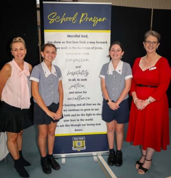Four people posed smilling with a banner displaying a school prayer. Two people are adults and two are children in school uniforms.