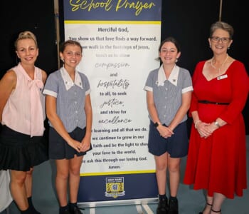 Four people posed smilling with a banner displaying a school prayer. Two people are adults and two are children in school uniforms.