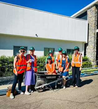Group of people in hard hats and high-viz gear posed smiling in front of a construction site, one person is sitting in a wheelbarrow.