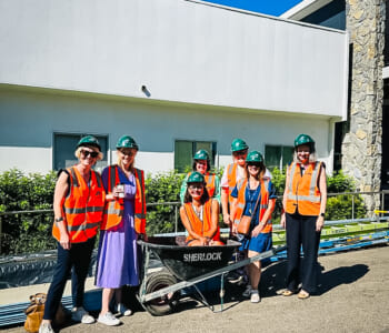 Group of people in hard hats and high-viz gear posed smiling in front of a construction site, one person is sitting in a wheelbarrow.
