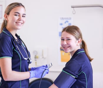 Two girls smiling in nurse uniforms one is holding a blood pressure measuring device and the other has the cuff around her arm