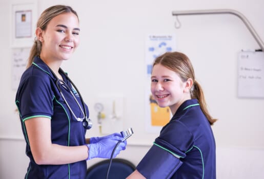 Two girls smiling in nurse uniforms one is holding a blood pressure measuring device and the other has the cuff around her arm