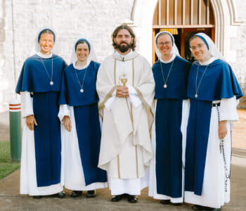 Group of smiling people posed together outside a Catholic church four are nuns in blue and white habits and one is a priest in white vestments