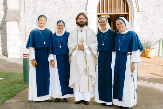 Group of smiling people posed together outside a Catholic church four are nuns in blue and white habits and one is a priest in white vestments