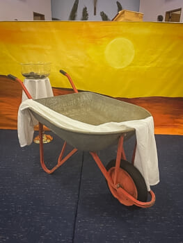 An art installation showing an empty wheelbarrow draped with a white cloth in front of a painted desert scene