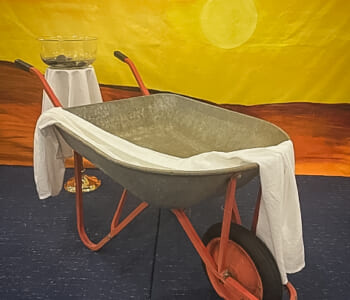 An art installation showing an empty wheelbarrow draped with a white cloth in front of a painted desert scene