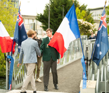 Two students shaking hands at the entrance to a bridge they are flanked by French and Australian flags