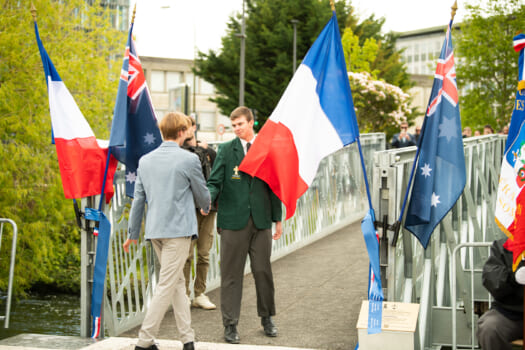 Two students shaking hands at the entrance to a bridge they are flanked by French and Australian flags