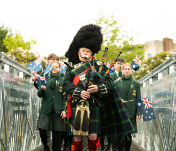 A bagpiper is leading a group of students across a bridge. The students are wearing green uniforms and waving Australian flags.