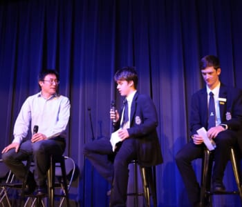 Three people sitting on stools talking one person is an adult and two a school students in uniform.