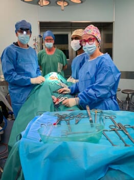 Four people performing surgery, they are wearing surgical masks and medical scrubs. The person being operated on is covered in a blanket and not visable.
