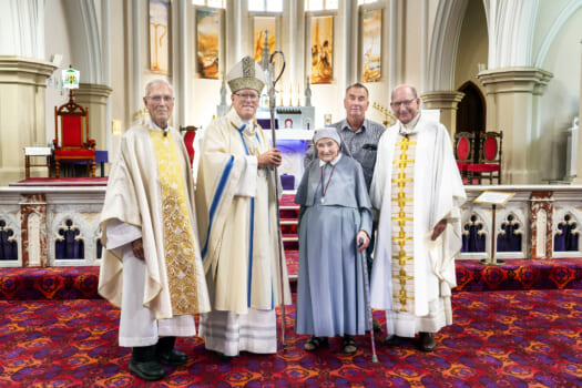 A group of people posed smiling within a church, three are men who are Catholic clergy, one is a nun in grey and the other is a gentleman in civilian clothes.