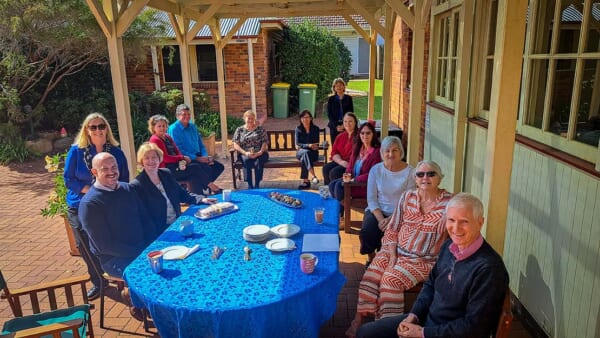 A group of people smiling gathered around a table sharing tea, coffee and snacks.